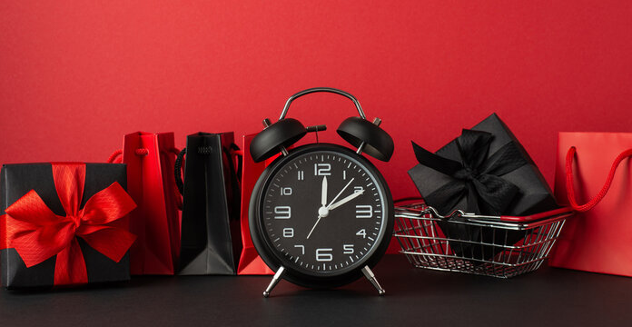 Black friday sales concept. Photo of alarm clock red and black paper bags and giftbox with ribbon bow in shopping cart on black desktop red wall background