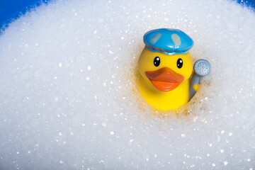 A yellow rubber duck for a bath in a bath cap and with a shower head in the wing in a cloud of soapy foam.
