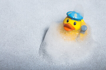 A yellow rubber duck for a bath in a bath cap and with a shower head in the wing in a cloud of soapy foam.