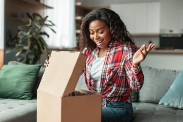 Shocked happy millennial african american woman opens cardboard box looks at purchases, enjoy sale