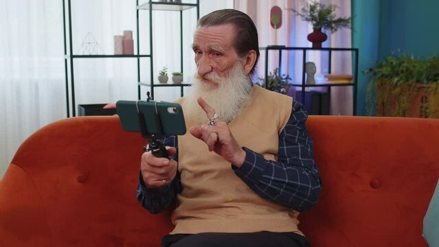 Senior old grandfather man blogger taking selfie on smartphone tripod, communicating, record video call online with subscribers. Elderly grandpa at modern home apartment living room sitting on couch