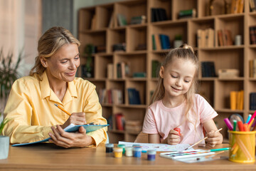 Happy female kid enjoying painting sitting with female child development specialist assisting cute little girl