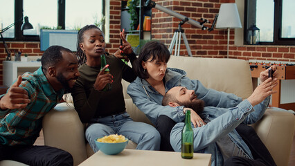 Multiethnic group of friends taking funny pictures on smartphone, making memories together and laughing at home party. Drinking beer and using phone to take photos, fooling around.