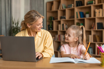 Fototapeta na wymiar Happy mother and her little daughter spending time together, cute girl drawing while her mom working on laptop