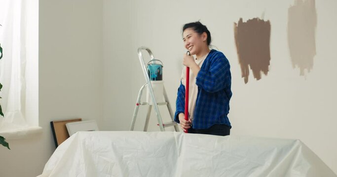 The Asian beauty is pleased with the progress of the renovation of new apartment. The girl paints the walls with a roller sings as she fools around dancing into a microphone.