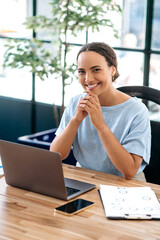 Vertical photo of a happy elegant pretty brazilian or latino woman, office employee, product or financial manager, sits at work desk in a modern office, looking at camera, smiling friendly