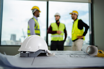 Selective focus of white helmet hardhat with a blueprint on the desk have a team of engineers...