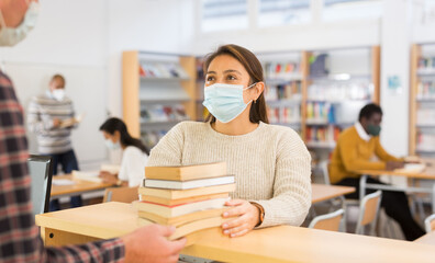 Portrait of young adult woman wearing face mask for viral protection returning books to librarian...