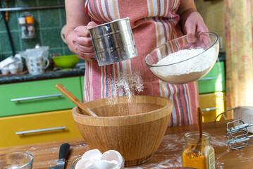 Hands of woman in apron sieving flour for homemade pastry over wooden bowl while standing by kitchen table.