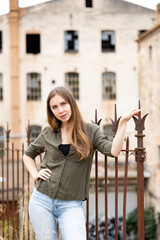 Naklejka na ściany i meble Gorgeous female model with long hair, dressed in jeans and shirt, posing against wrought-iron fence and building on background