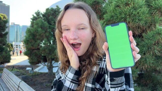 A young woman sits with a phone turned to the camera with a green screen. She sits in the center of Vancouver Porto Canada Place Coniferous trees are in the background. She is wearing checkered blouse