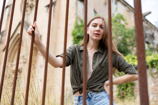 Attractive woman in jeans and shirt posing on camera on street. Female model with blond hair looking to camera and standing near wrought-iron fence