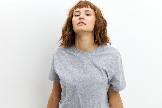 a sweet, beautiful, red-haired, curly-haired woman stands on a white background in a gray cotton T-shirt and looks at the camera with a neutral gaze. Horizontal studio photo with an empty space 