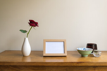 Small blank picture frame on oak side table with single red rose and bowls against beige wall