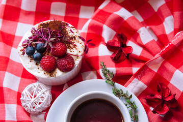 dessert with raspberries and blueberries in a ramekin with tea in a white cup and saucer on a red checkered napkin top view