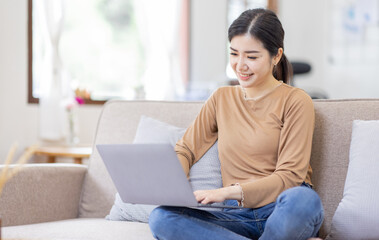 Beautiful Young Asian woman using laptop computer on sofa at home, shopping online with internet in happy mood and Relax part time concept