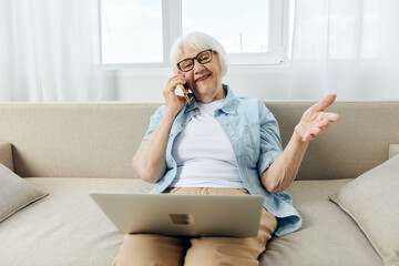 Fototapeta a happy lady in glasses and a light shirt is sitting in her apartment working remotely and holding her hand in a relaxed gesture talking on the phone holding a laptop on her lap obraz