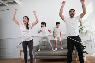 Happy wellness Asian Thai family, parents, and children are fun playing hula hoops together, fitness training and healthy exercise in white living room, domestic home lifestyle, and weekend activity.