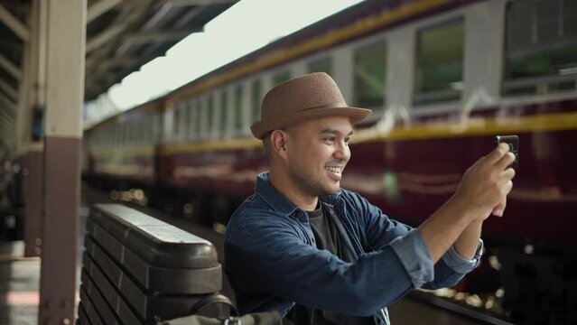 Happy tourist travel by train on vacation time holiday weekend trip. Freedom traveler young asian man at terminal train station and taking photo by smartphone. Backpacker arrival at platform railway.