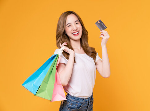 Beautiful Asian woman holding multi colored shopping bags and showing credit card on yellow background.