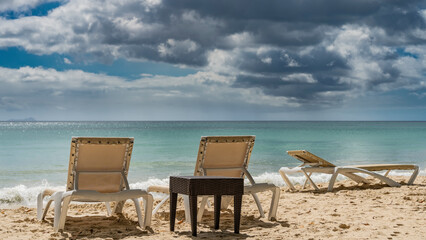 Sun loungers and a wicker table stand on the sandy beach. The turquoise ocean is calm. Foam of waves on the shore. Picturesque clouds in the blue sky. Seychelles. Mahe. Beau Vallon