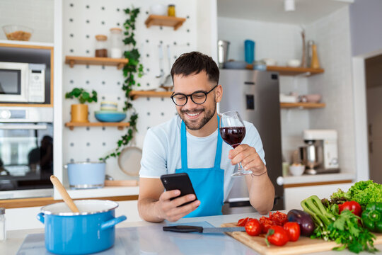 Head shot smiling young man using smartphone, chatting in social network while preparing food for vegetarian dinner at home. Happy millennial guy in eyeglasses web surfing recipe for meal in kitchen.