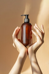 Bottle with dispenser, sprayer, for liquid soap, shampoo, lotion in women's hands on beige background. Rays of sunlight fall on container made of amber glass. Mockup concept, vertical frame