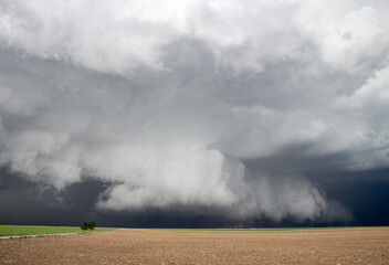 Light shines on a thick shelf cloud hanging low to the ground under a severe storm in farmland.