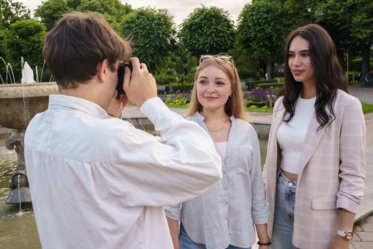 Portrait of young guy photographing his two female friends around park fountain.