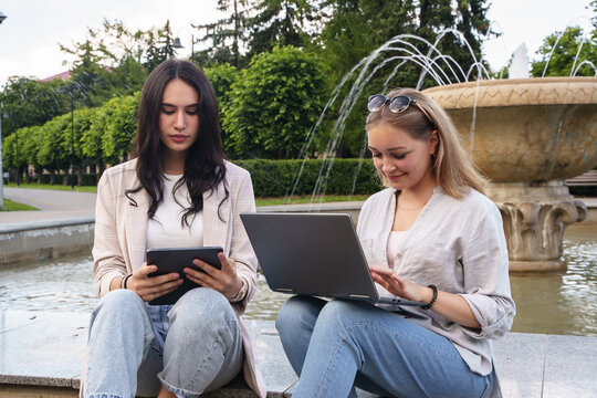 Shot of two female friends browsing tablet and laptop sitting by fountain in park.