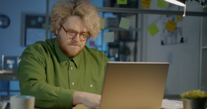 Determined office worker is using laptop late at night typing and looking at screen with crazy eyes. Overworker young people and aspirations concept.