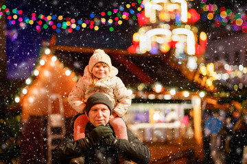Little preschool girl sitting on shoulder of father on Christmas market in Germany. Happy toddler child and man observing traditional decorated pyramid. Happy family, bonding, love. Family xmas time.