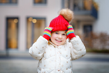 Funny portrait of little preschool girl in winter clothes. Happy positive child with red hat and colorful gloves outdoors. Winter day.