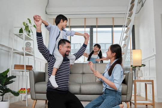 Asian Thai Family Together, Dad Plays And Teases With Children And Mum By Carrying And Holding Son On Shoulders In Home Living Room, Happy Leisure Times, Lovely Weekend, Wellbeing Domestic Lifestyle.