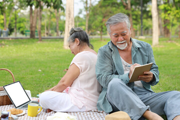 Fototapeta na wymiar Happy asian senior man and woman sitting on blanket and having fun on picnic together in garden outdoor. Lover couple using computer and writing at the park. Happiness marriage lifestyle concept.
