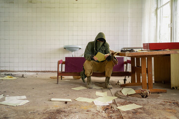 Man in bandana mask, hood and camo pants sits inside of abandoned building and reading documents. Cyberpunk postapocalypse fantasy horror scene,