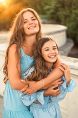 Fototapeta premium Portrait of mother and daughter in blue dresses with flowing long hair against the backdrop of sunset. The woman hugs and presses the girl to her. They are looking at the camera.