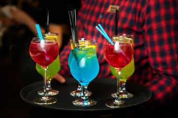 The waiter holds a tray with Multicolored alcoholic and non-alcoholic cocktails with ice and straws