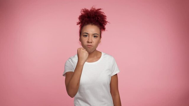 Portrait serious African American redhead curly woman threatens with shaking fist looking camera. Female in white t-shirt standing on isolate pink background with copy space