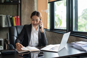 Fototapeta premium Happy young Asian woman talking on the mobile phone and smiling while sitting at her working place in an office.