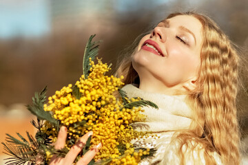 Naklejka premium Portrait of a young woman with blond hair holding a bouquet of mimosa in her hands. Spring