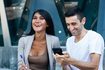 Smiling multiracial couple looking at cellphone outside office building during work break. Young man showing digital document to Thai woman colleague, teamwork, white collar job concepts