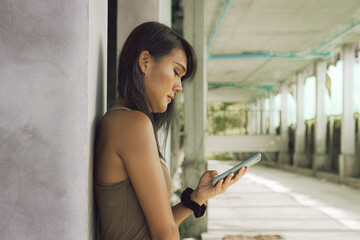 Lonely Thai woman sending text message by cellphone leaning on concrete column of abandoned building. Transgender person from the side using mobile phone