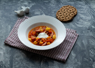 Fototapeta Soup with sausages and sour cream in a white plate on a striped napkin. Two slices of bread. obraz
