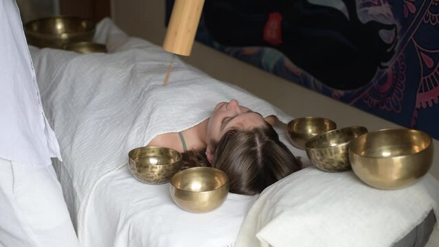 A man wearing in white using a bamboo Koshi chime musical instrument during sound healing therapy with tibetan singing bowls over young woman during sound massage indoor, selective focus