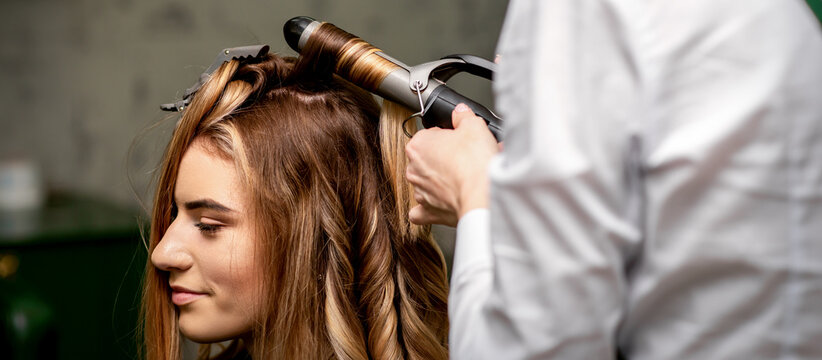 The female hairdresser is curling hair for a brown-haired young caucasian woman in a beauty salon