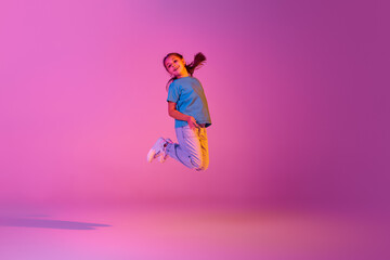 Fototapeta na wymiar Beautiful active little girl, kid jumping, dancing isolated over pink background in neon. Action, dance, happy childhood