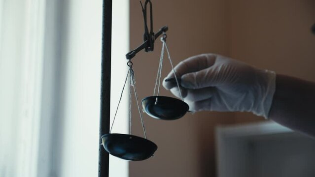 Close-up of medical worker hand in rubber gloves measuring medicine, putting weight on traditional metal balance scale, scales moving due imbalance. Concept of weights comparison.