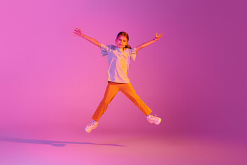 Fototapeta na wymiar Expressive little girl, kid in casual bright clothes jumping isolated over pink background in neon. Action, dance, happy childhood