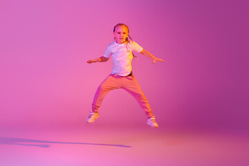 Fototapeta na wymiar Beautiful active little girl, kid jumping, dancing isolated over pink background in neon. Action, dance, happy childhood